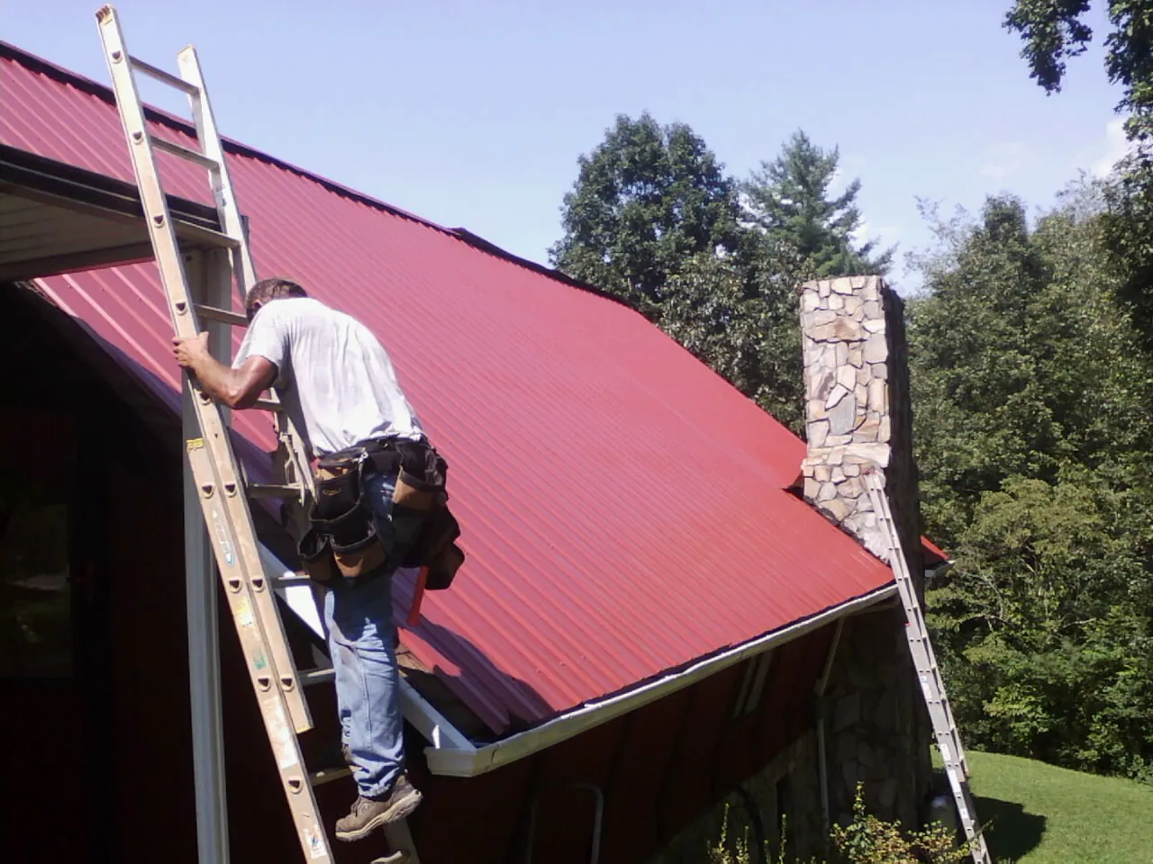 Roofer on a ladder beside a red metal roof and stone chimney — Soffit & Fascia Repair by Roofing Brothers in Blountsville